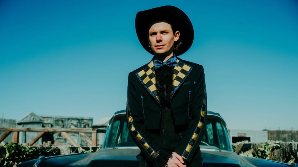 A photograph of Hayden Pedigo standing in front of a car wearing a suit and hat.